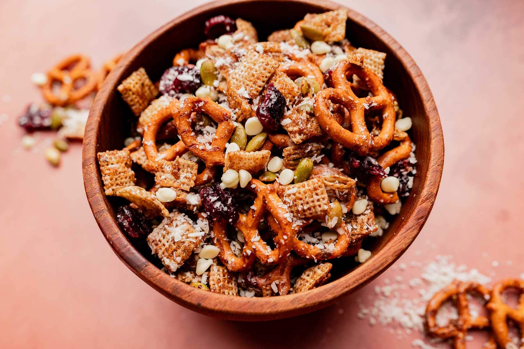 A wooden bowl filled with christmas snack mix—pretzels, Chex cereal, dried cranberries, white chocolate chips, seeds, and coconut flakes—with some of the festive treat spilled on the surface.