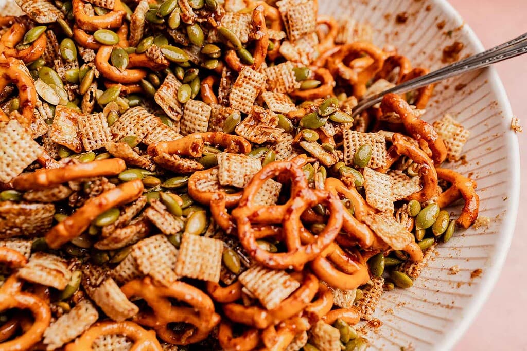 A close-up of a bowl filled with pretzels, cereal squares, and pumpkin seeds makes for a festive Christmas snack mix, with a metal spoon partially visible on the side.
