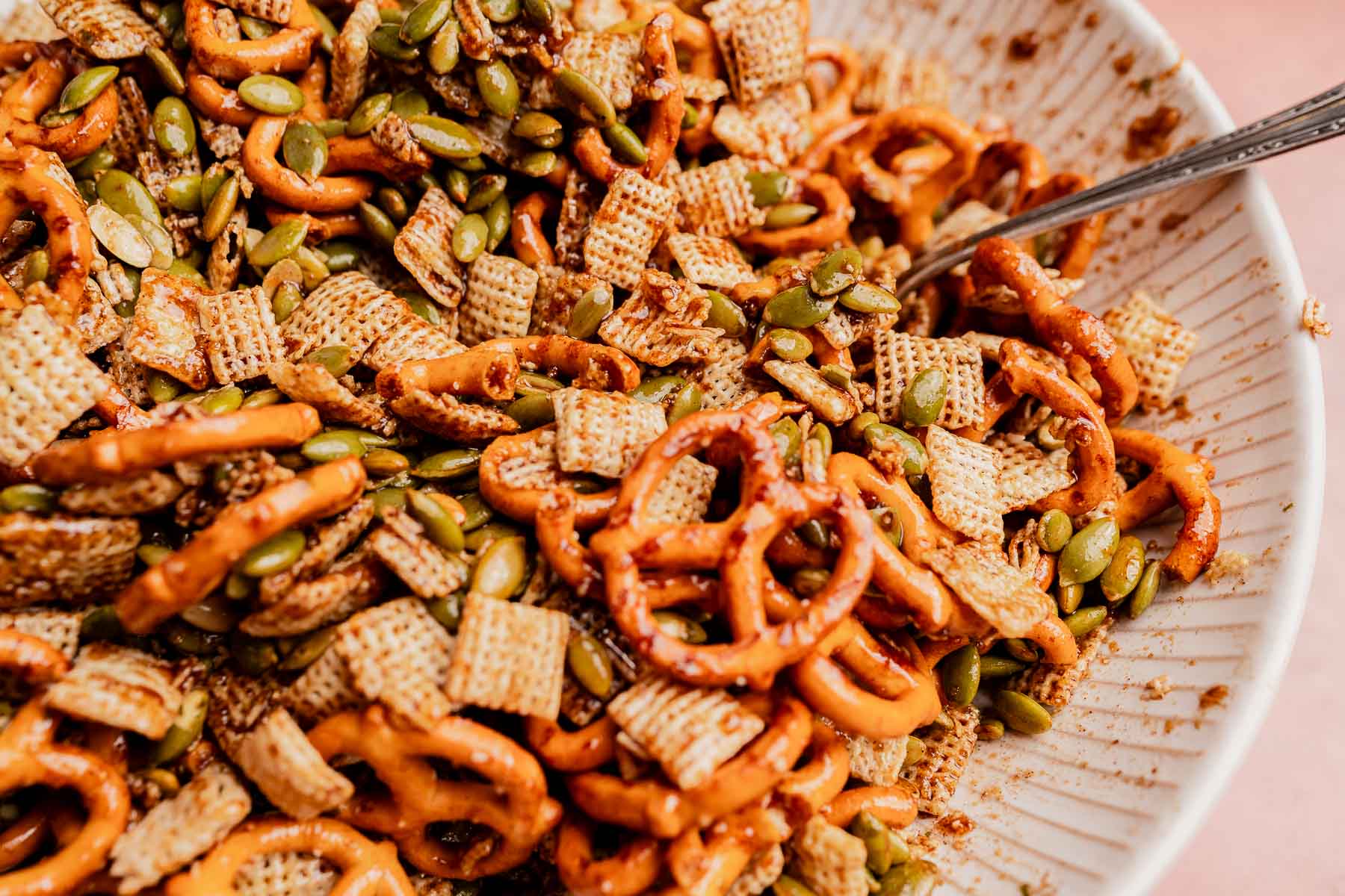 A close-up of a bowl filled with pretzels, cereal squares, and pumpkin seeds makes for a festive Christmas snack mix, with a metal spoon partially visible on the side.