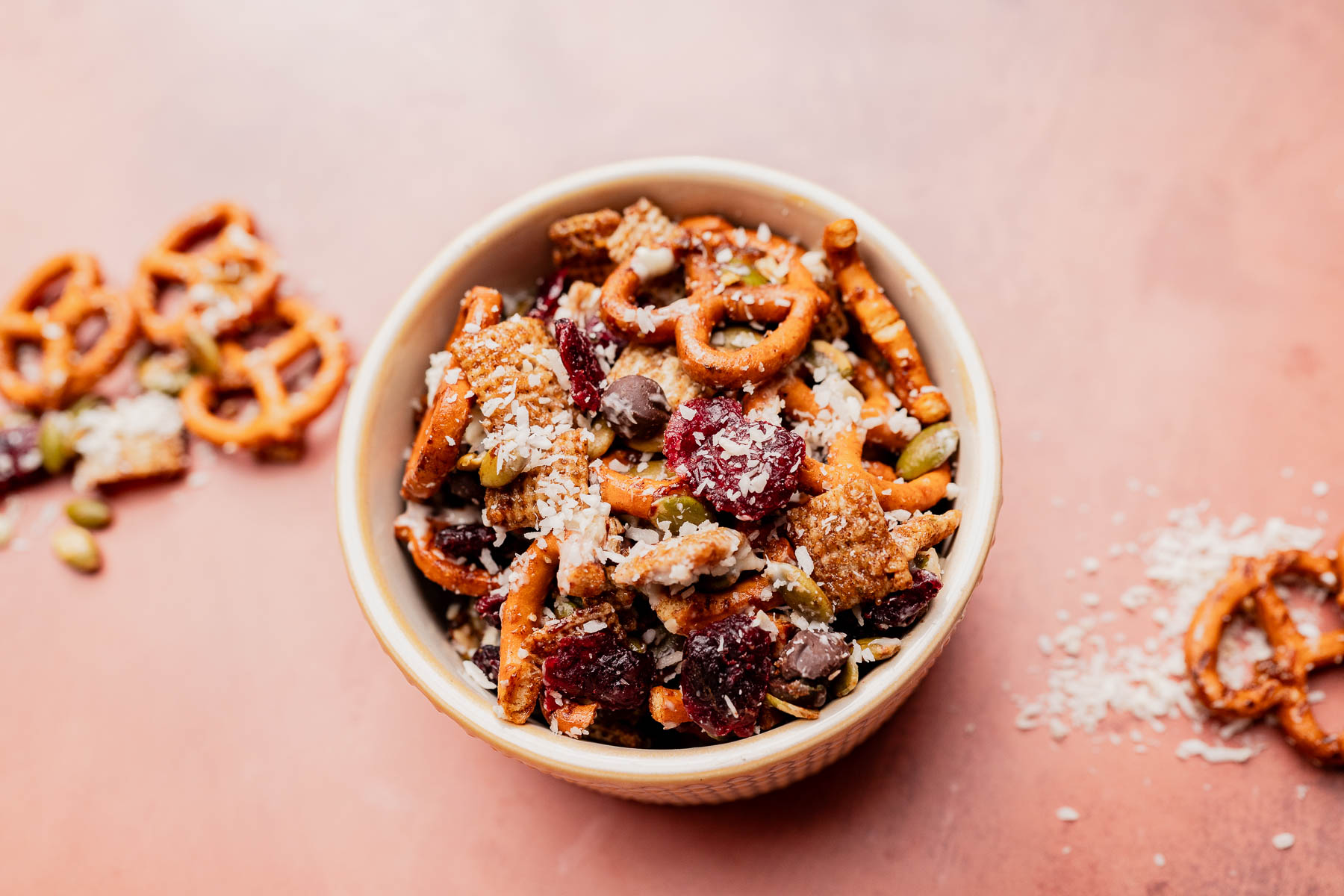 A bowl of christmas snack mix featuring pretzels, dried fruit, chocolate chips, seeds, and shredded coconut sits on a pink surface.