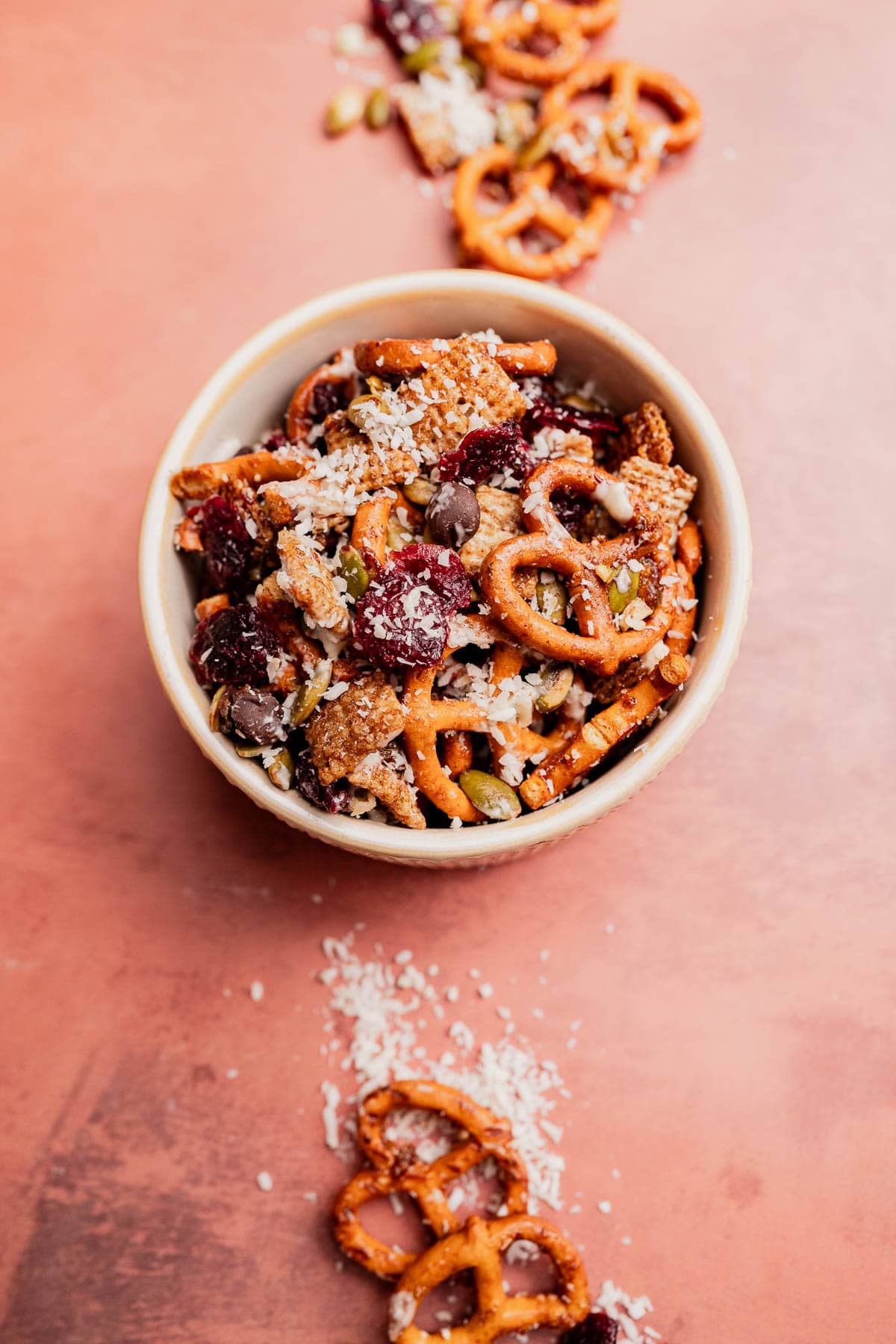 A bowl of Christmas snack mix with pretzels, dried cranberries, chocolate chips, seeds, and shredded coconut sits on a pink surface, with some festive mix scattered outside the bowl.
