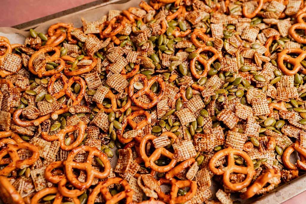 A tray of christmas snack mix featuring pretzels, Chex cereal, and pumpkin seeds spread out on parchment paper.