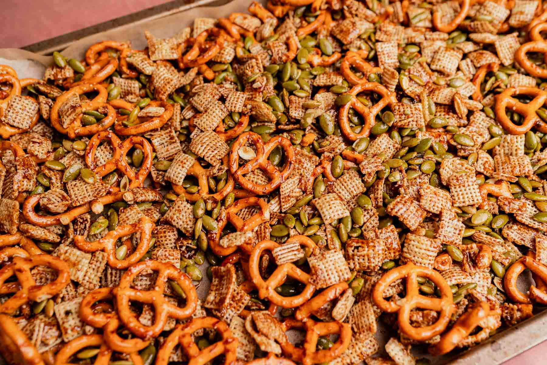 A tray of christmas snack mix featuring pretzels, Chex cereal, and pumpkin seeds spread out on parchment paper.
