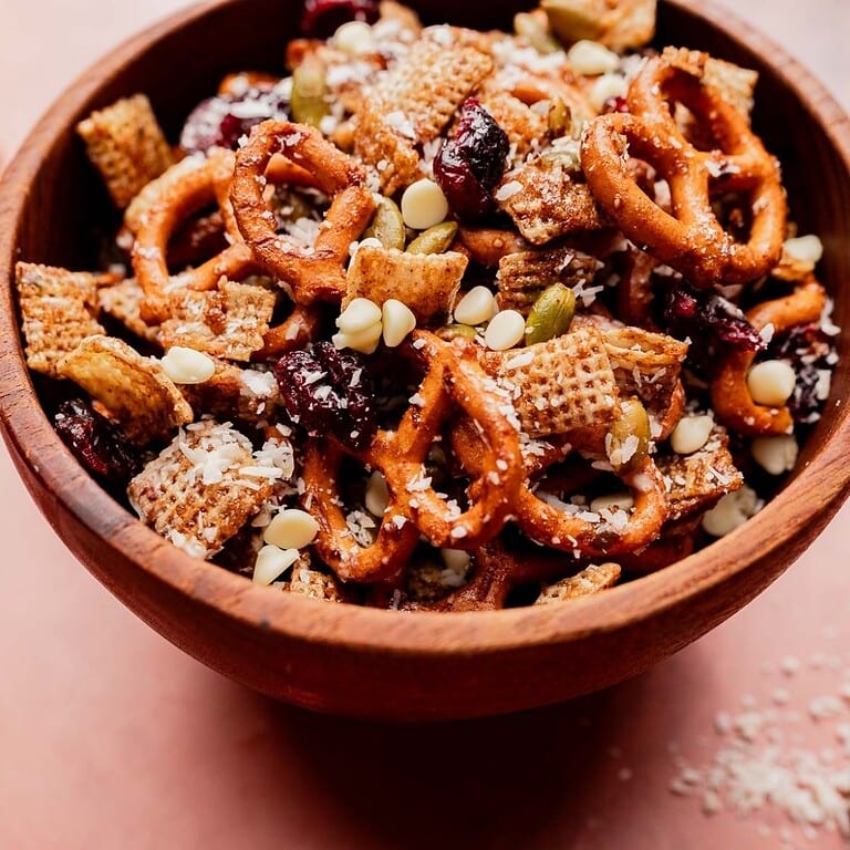 A wooden bowl filled with a festive Christmas snack mix of pretzels, cereal squares, white chocolate chips, dried cranberries, pumpkin seeds, and shredded coconut.