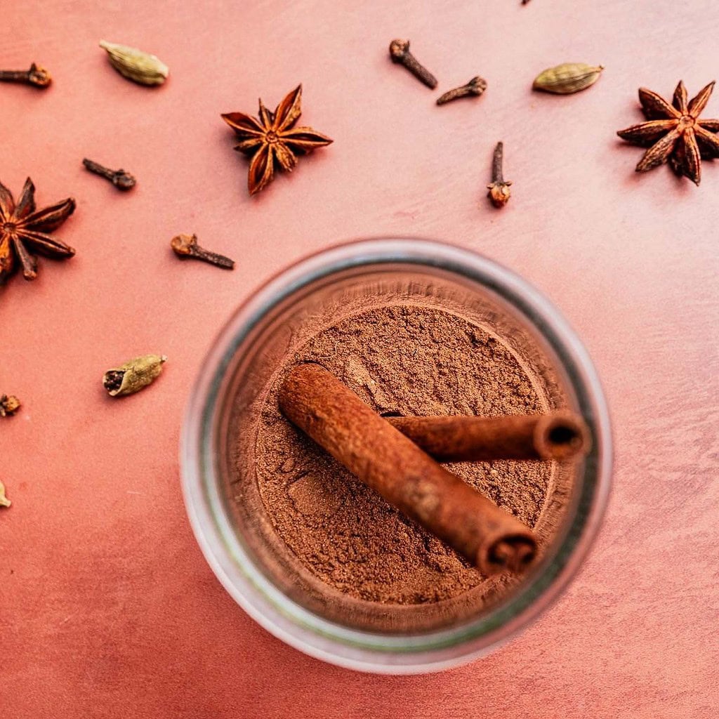 A jar of ground christmas spice blend with two cinnamon sticks inside, surrounded by star anise, cardamom pods, and cloves on a pink surface.