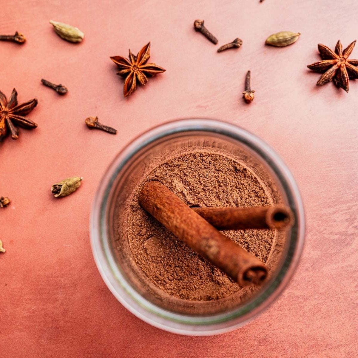 A jar of ground christmas spice blend with two cinnamon sticks inside, surrounded by star anise, cardamom pods, and cloves on a pink surface.