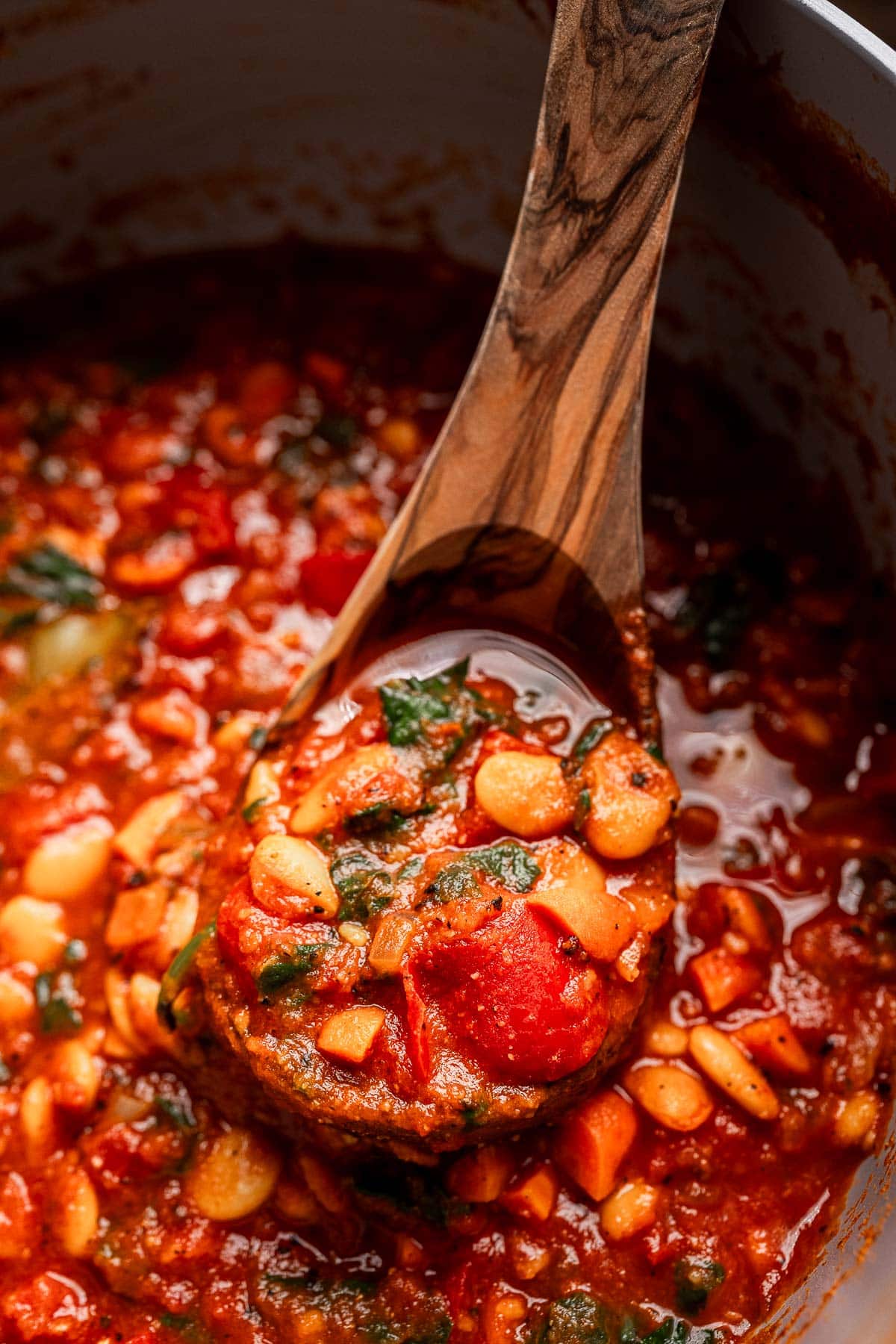 A wooden spoon scoops hearty butter bean stew, brimming with white beans, tomatoes, and greens, from a pot.