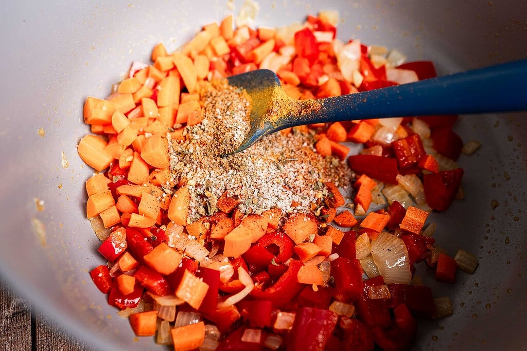 Chopped carrots, onions, and red bell peppers are sautéed in a pan to form the flavorful base of a hearty butter bean stew, with dry seasoning stirred in using a blue spatula.