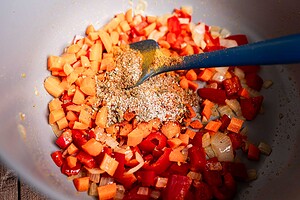 Chopped carrots, onions, and red bell peppers are sautéed in a pan to form the flavorful base of a hearty butter bean stew, with dry seasoning stirred in using a blue spatula.