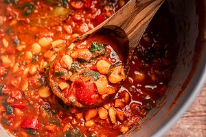 A wooden spoon lifts a portion of butter bean stew, rich with tomatoes, herbs, and vegetables, from a pot.
