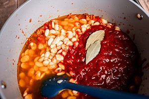 A close-up of butter bean stew with white beans, tomato sauce, chopped tomatoes, and bay leaves simmering in a pot, as a blue spatula stirs the hearty mixture.