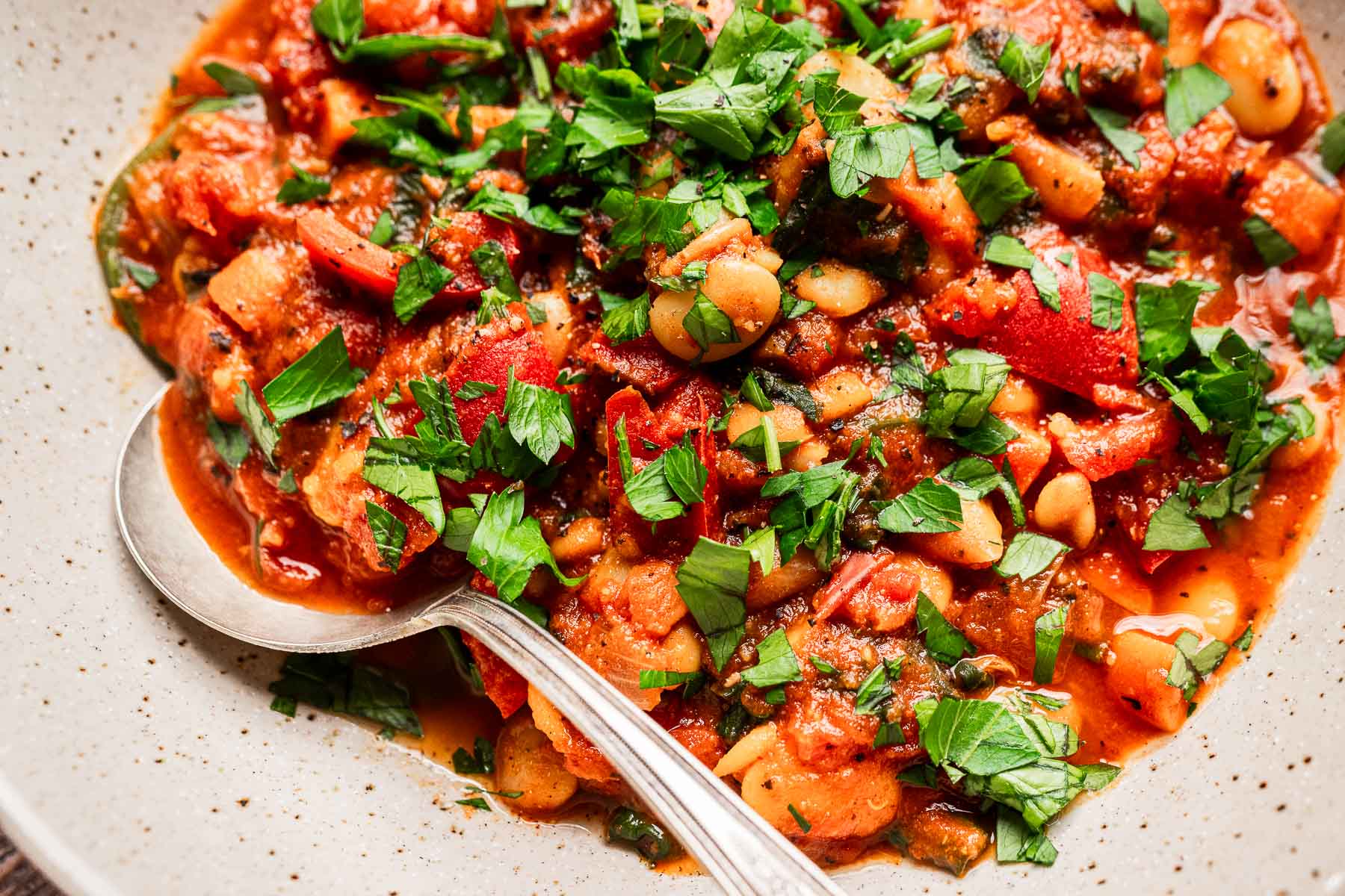 A close-up of a bowl of tomato-based vegetable and butter bean stew with white beans, garnished with chopped fresh parsley, and a spoon resting in the bowl.
