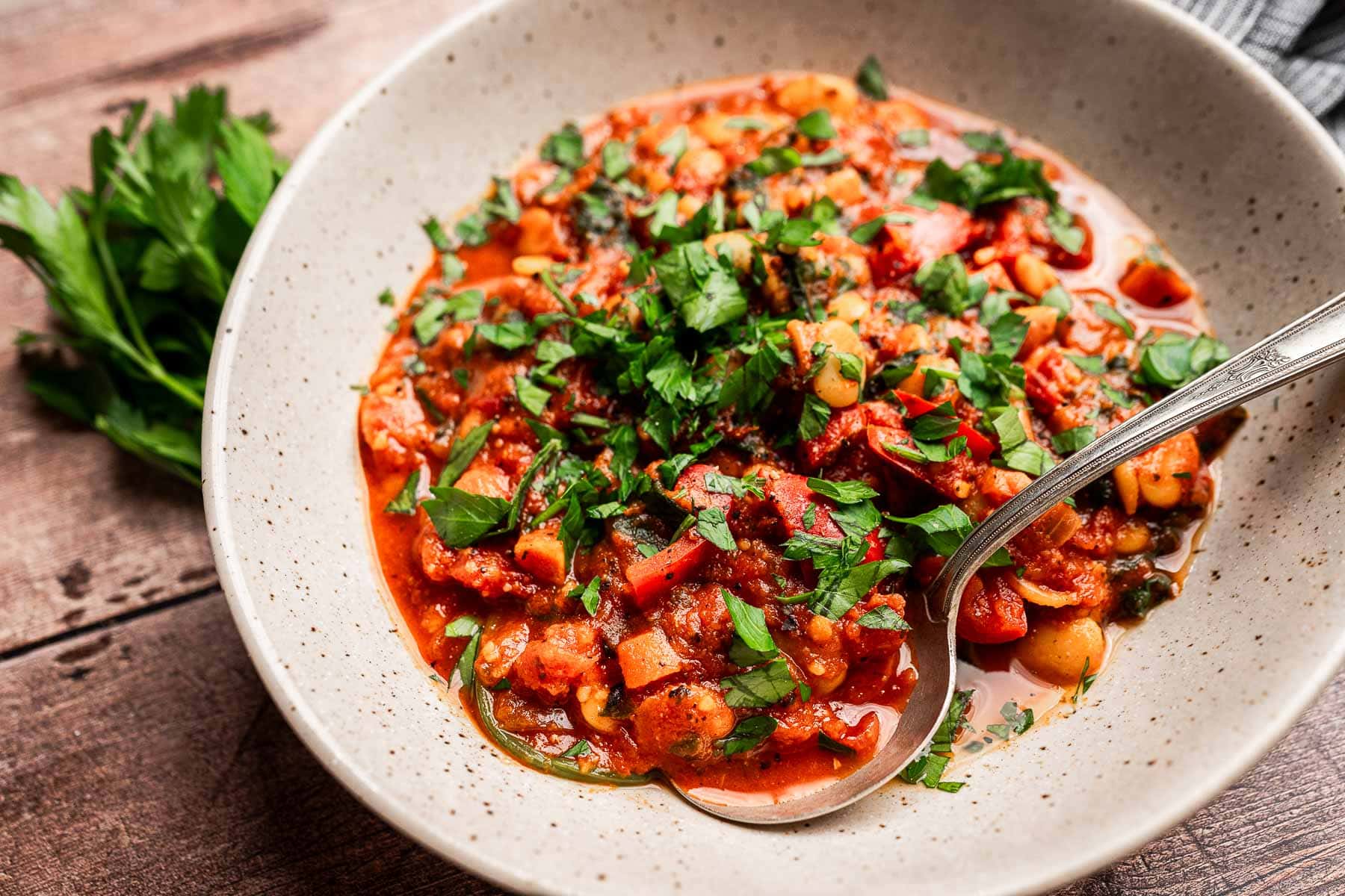 A bowl of butter bean stew with chickpeas, tomatoes, and herbs, garnished with chopped parsley, sits on a wooden table with a spoon in the bowl.