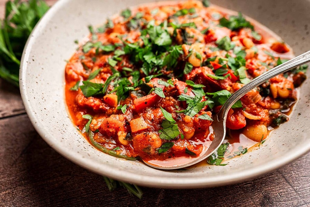 A bowl of butter bean stew with a tomato-based sauce, garnished with chopped fresh herbs, and a spoon resting inside.