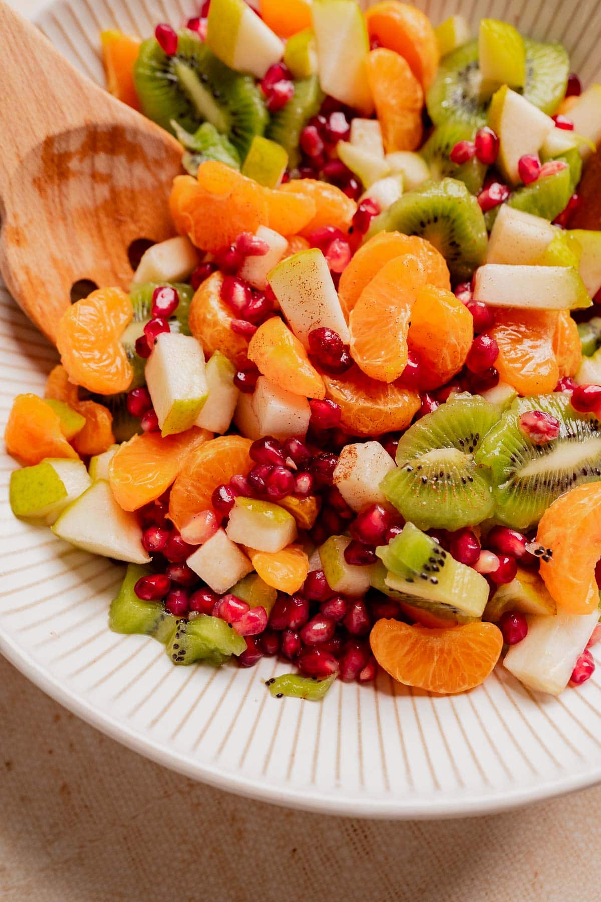 A bowl of christmas fruit salad featuring kiwi, mandarin orange segments, pear pieces, and pomegranate seeds, served with a wooden spoon.