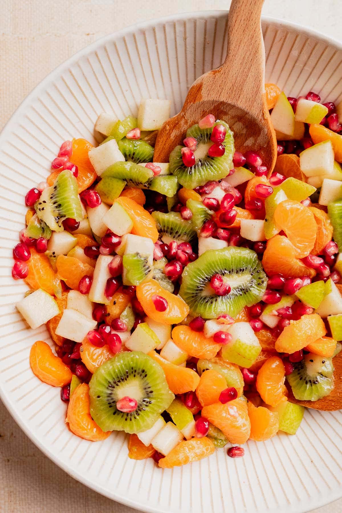 A festive bowl of Christmas fruit salad featuring sliced kiwi, mandarin orange segments, diced apple, and pomegranate seeds, all served with a wooden spoon.