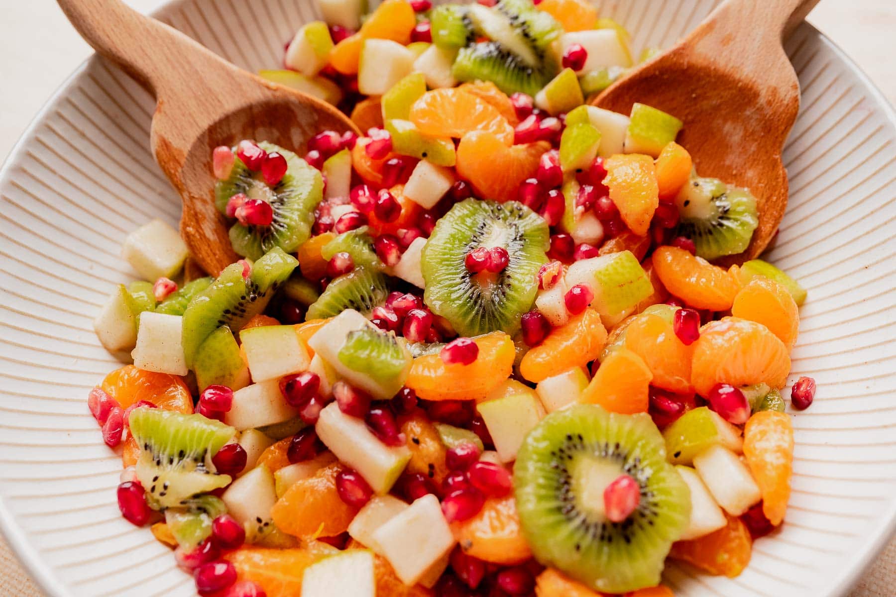 A bowl of christmas fruit salad with kiwi, mandarin oranges, apple, green apple, and pomegranate seeds is served with two wooden salad spoons.