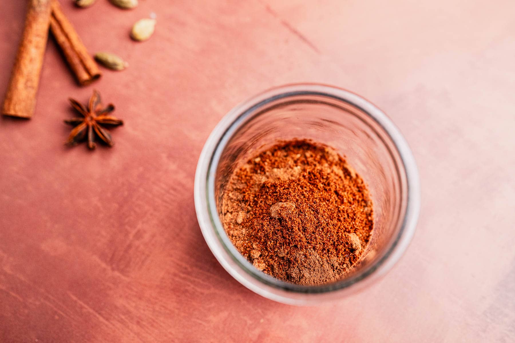 A glass jar filled with christmas spice blend sits on a pink surface, surrounded by whole spices like cinnamon sticks, cardamom pods, and star anise.