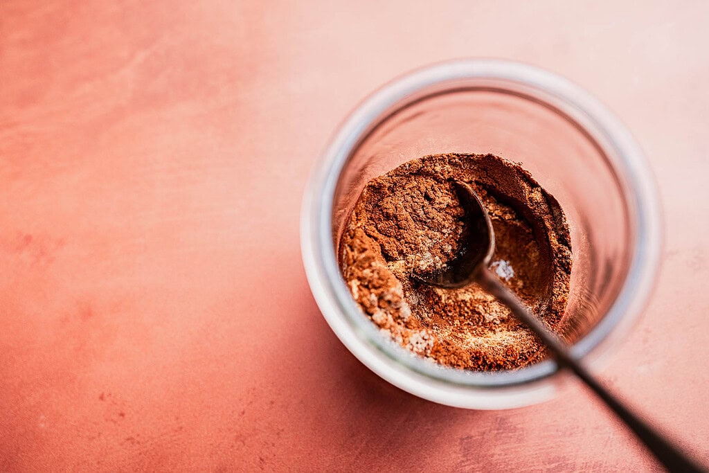A glass jar containing a Christmas spice blend of brown spices with a black spoon inside, placed on a peach-colored surface.