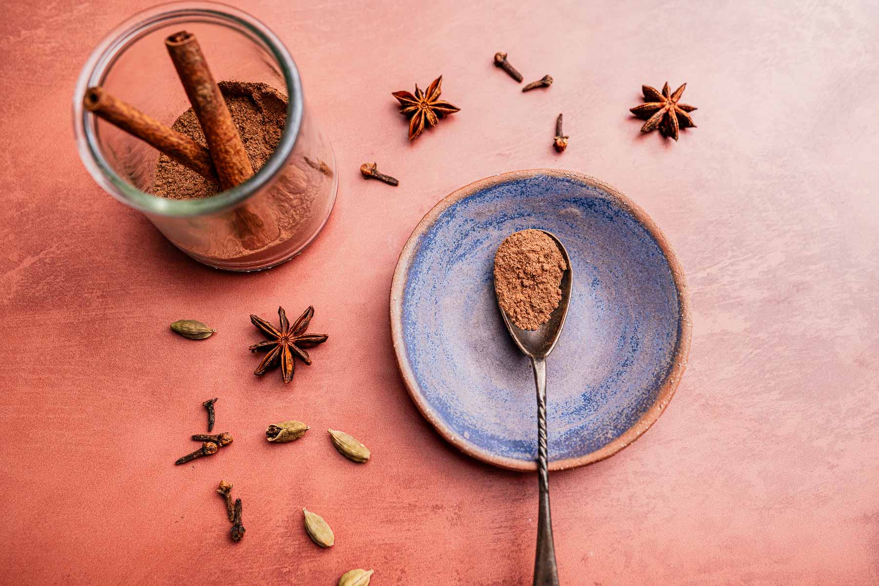 A jar of christmas spice blend with cinnamon sticks, a blue plate holding a spoonful of the blend, and scattered star anise, cardamom pods, and cloves on a pink surface.