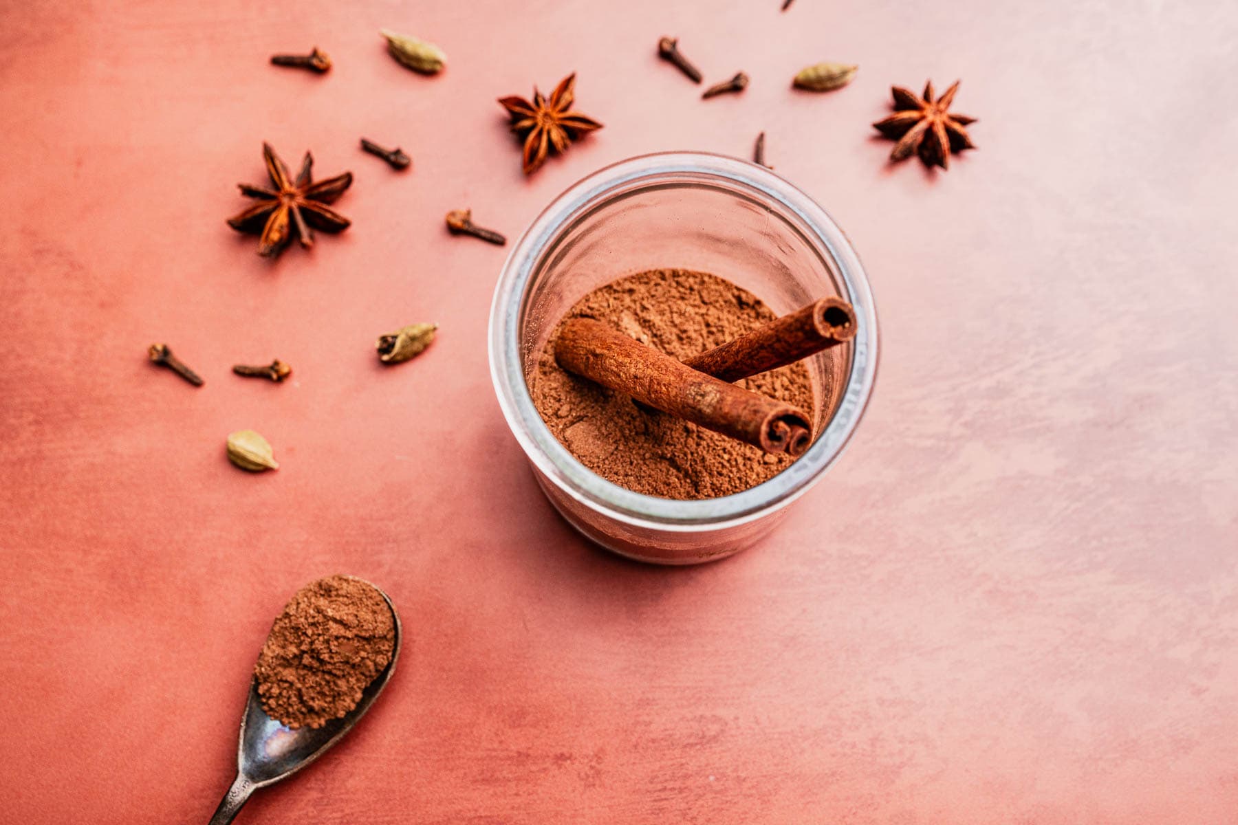 A glass jar with christmas spice blend, topped with two cinnamon sticks, is surrounded by whole spices like cloves, cardamom, and star anise on a pink surface. A spoon holds ground spice.