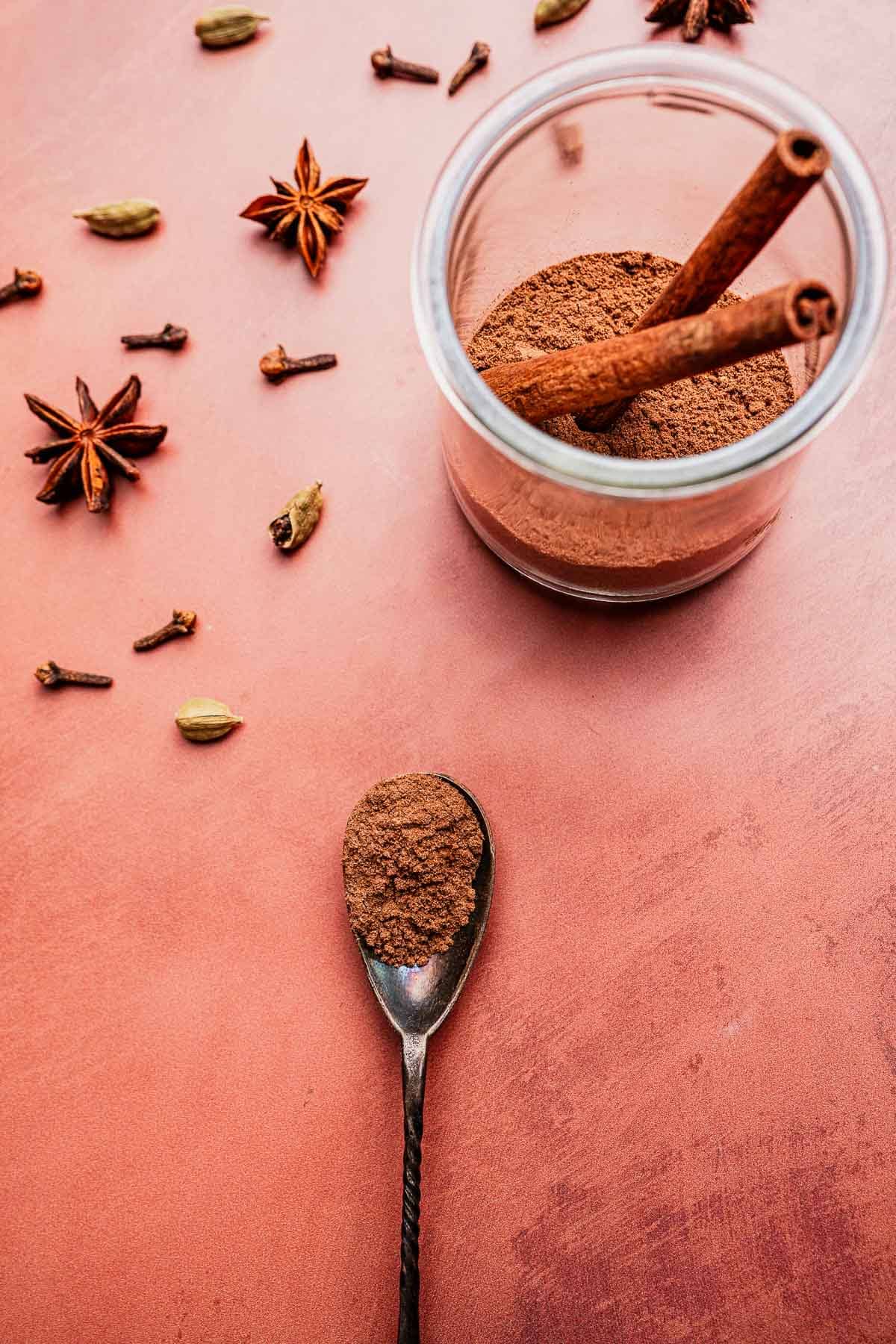 A spoon with ground spice, a jar of powdered christmas spice blend with cinnamon sticks, and whole spices including star anise, cloves, and cardamom on a pink surface.