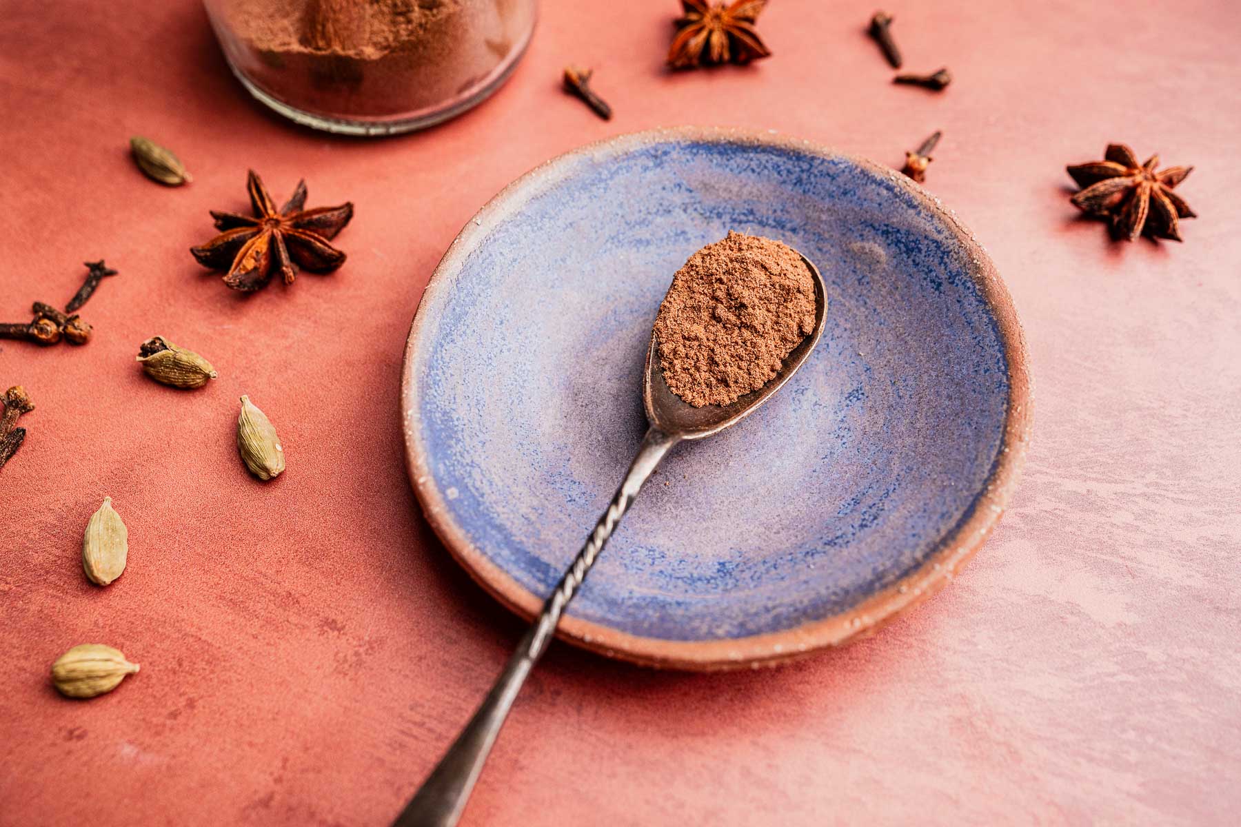 A spoonful of christmas spice blend powder rests on a blue ceramic plate, surrounded by whole star anise, cloves, and cardamom pods on a pink surface.