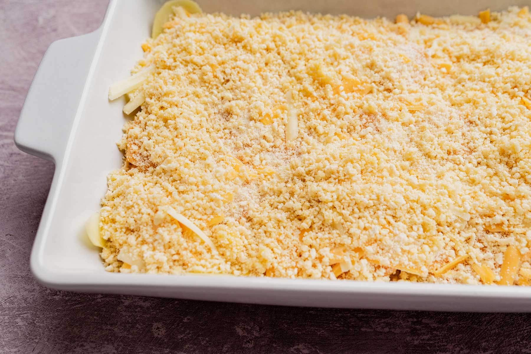 A white baking dish filled with an unbaked macaroni and cheese casserole, reminiscent of a classic potato casserole, topped with a layer of breadcrumbs and shredded cheese.