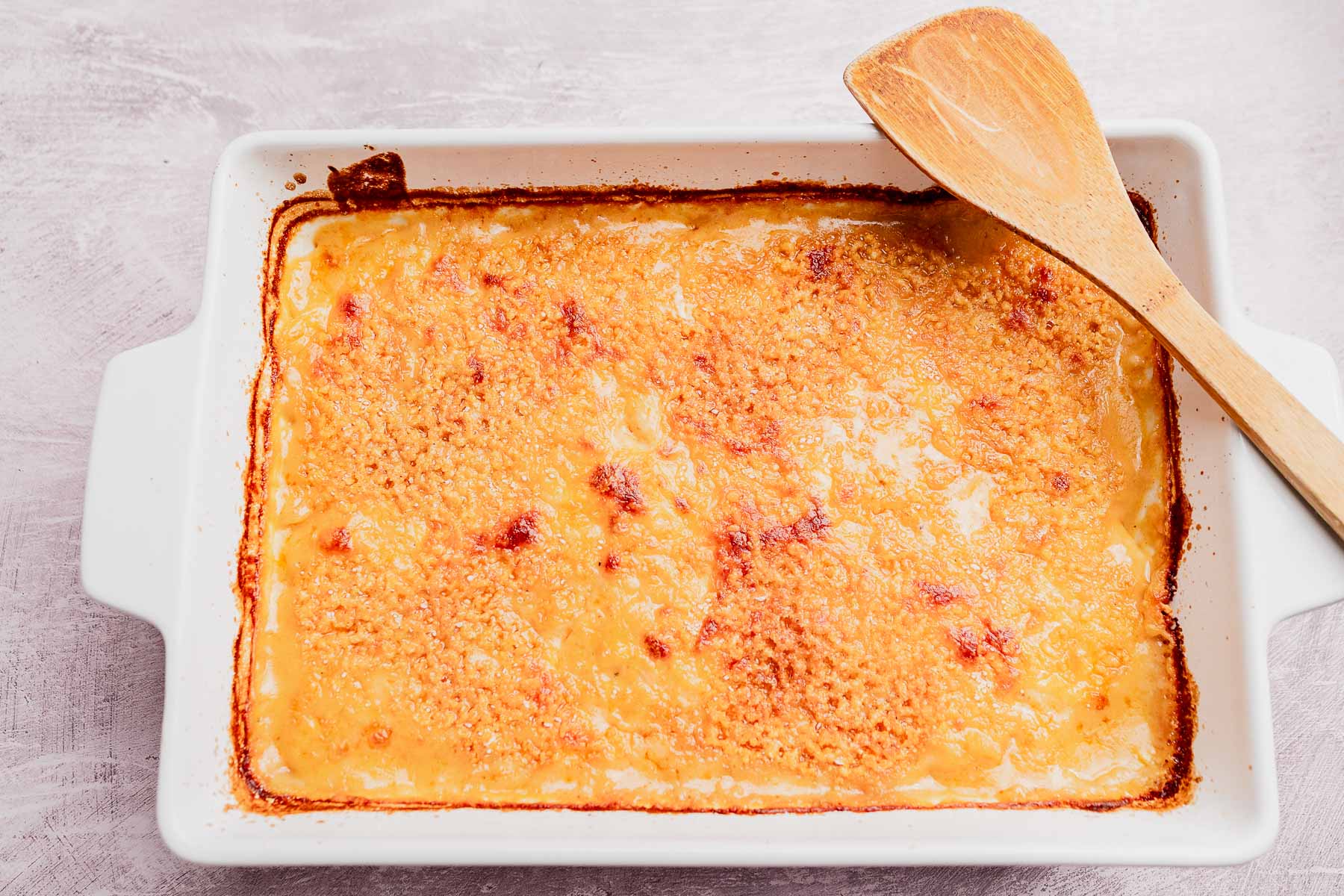 A rectangular baking dish filled with baked macaroni and cheese or potato casserole, both topped with golden breadcrumbs, with a wooden spoon resting on the edge.