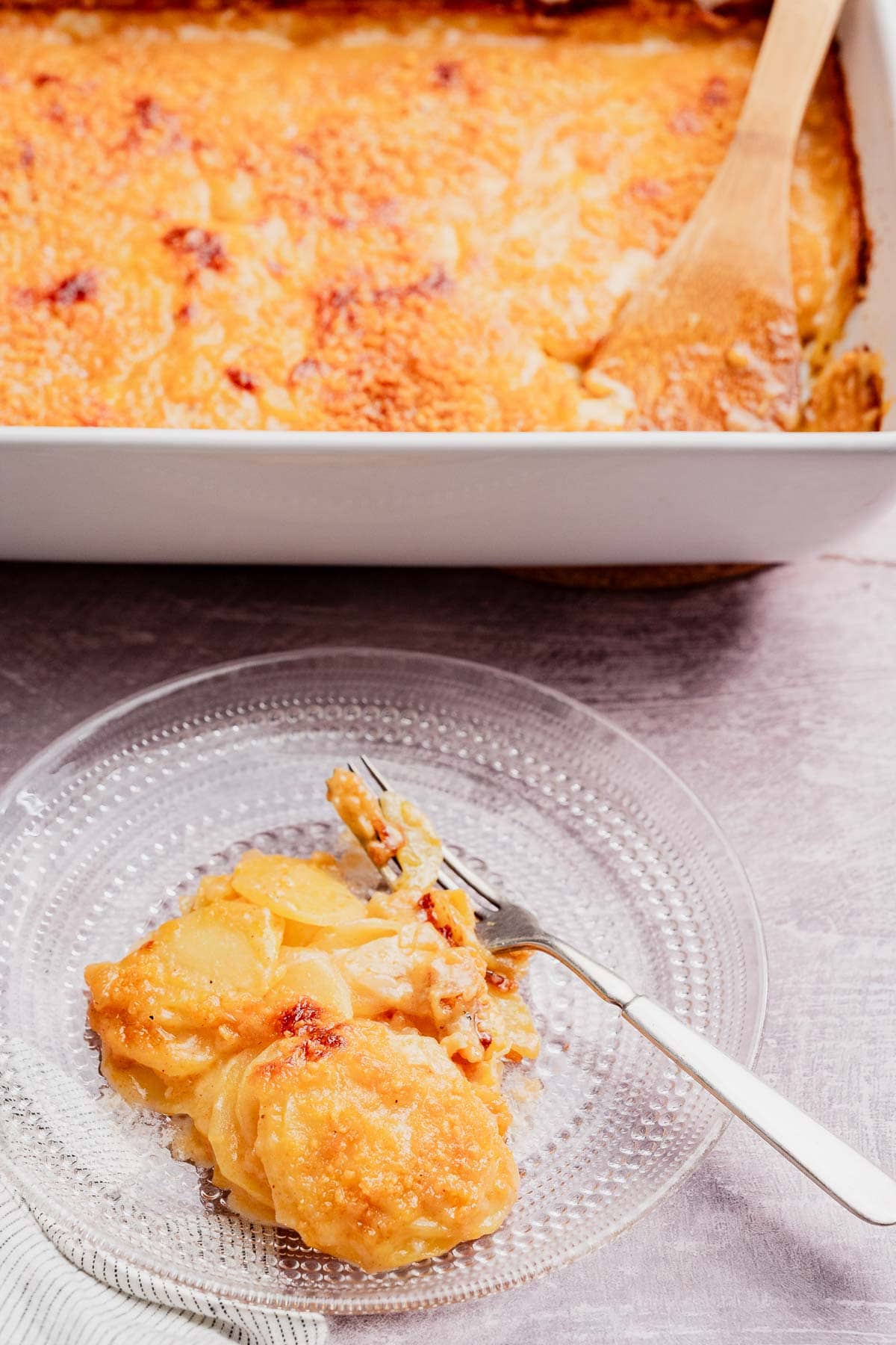 A serving of baked potato casserole on a clear glass plate with a fork, in front of a casserole dish filled with the remaining potatoes and a wooden spoon.