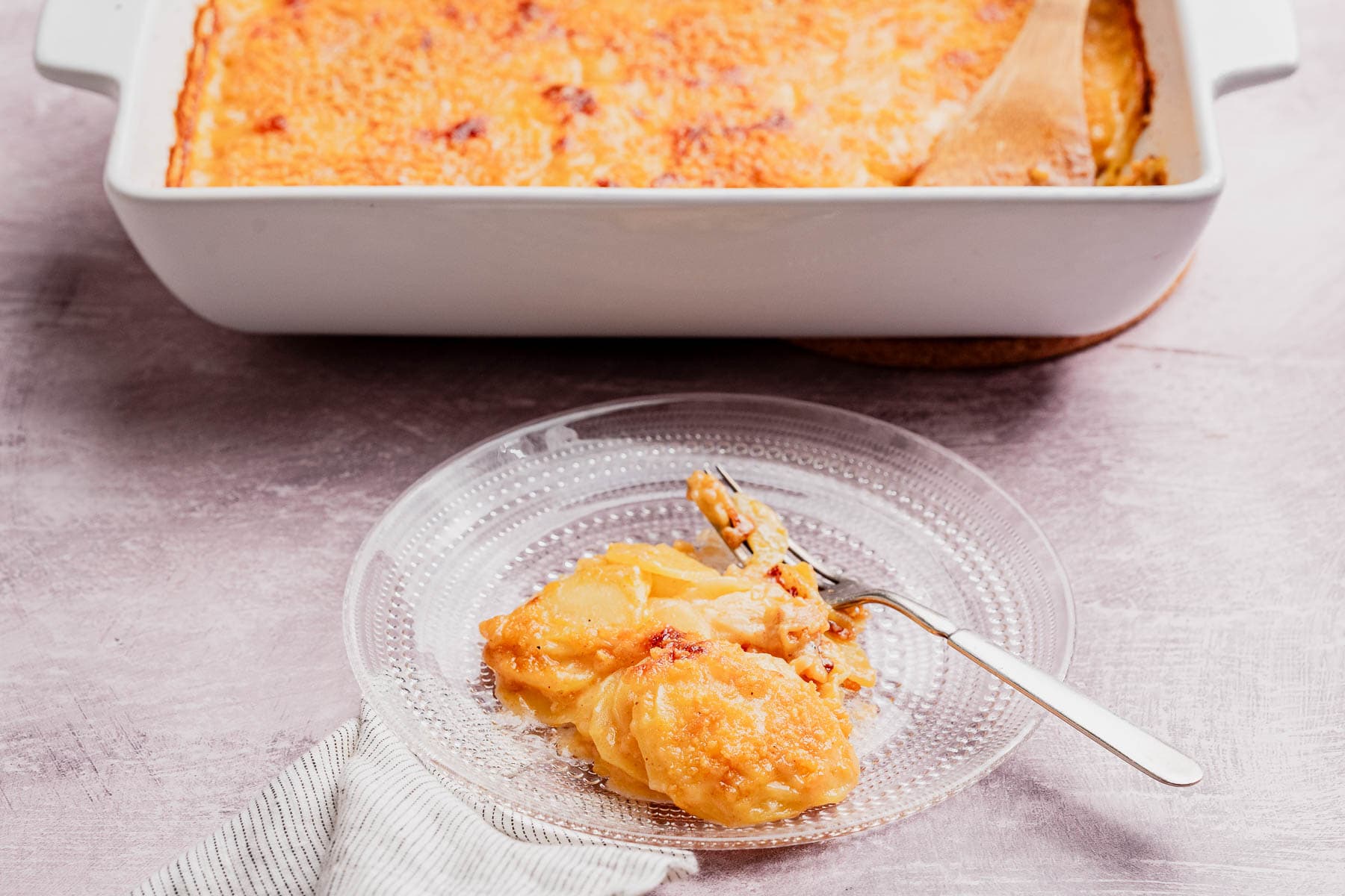 A clear glass plate holds a serving of cheesy potato casserole gratin next to a fork, set in front of a baking dish filled with more gratin and a wooden spoon.
