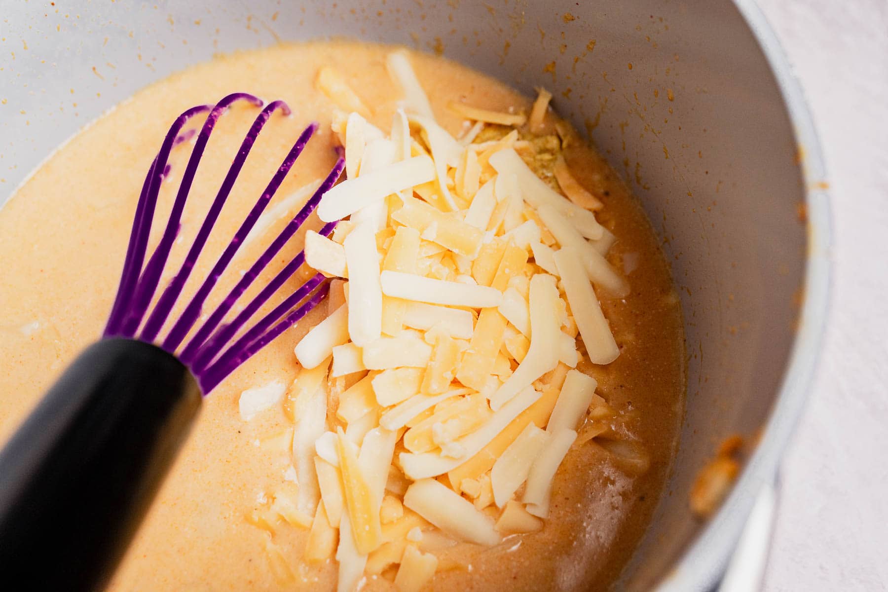 A purple whisk sits in a pot of creamy orange sauce, perfect for a potato casserole, with shredded cheese on top, partially melted.