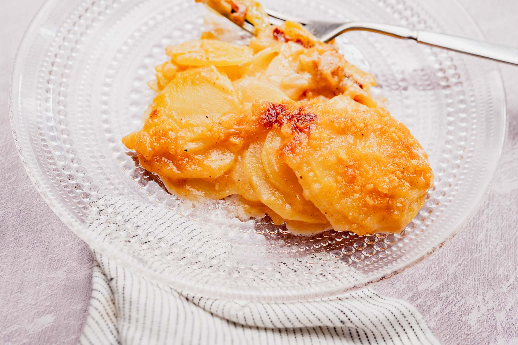 A serving of cheesy potato casserole sits on a clear glass plate, with a fork and striped napkin beside it.