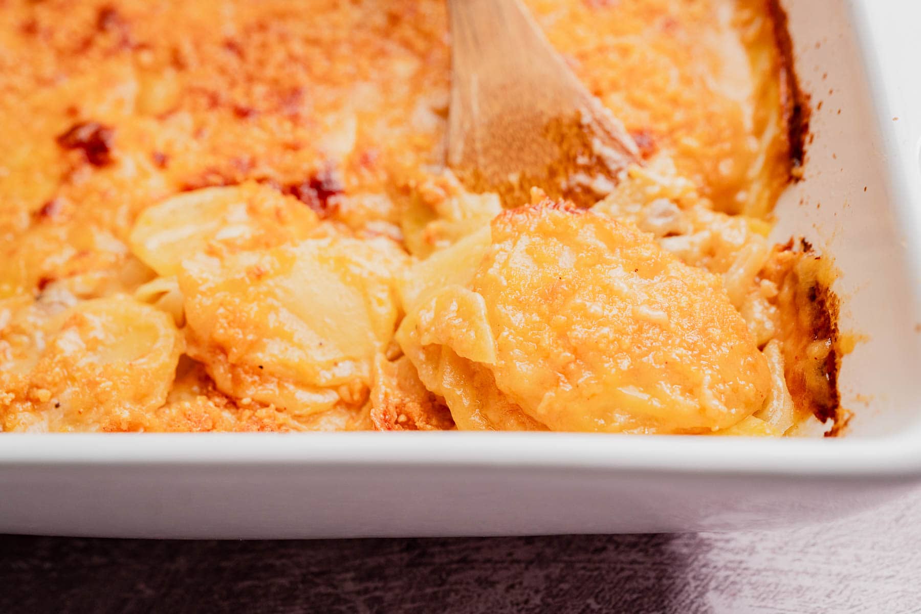 A close-up of a baking dish filled with cheesy scalloped potatoes, this comforting potato casserole is shown as a wooden spoon scoops out a hearty portion.