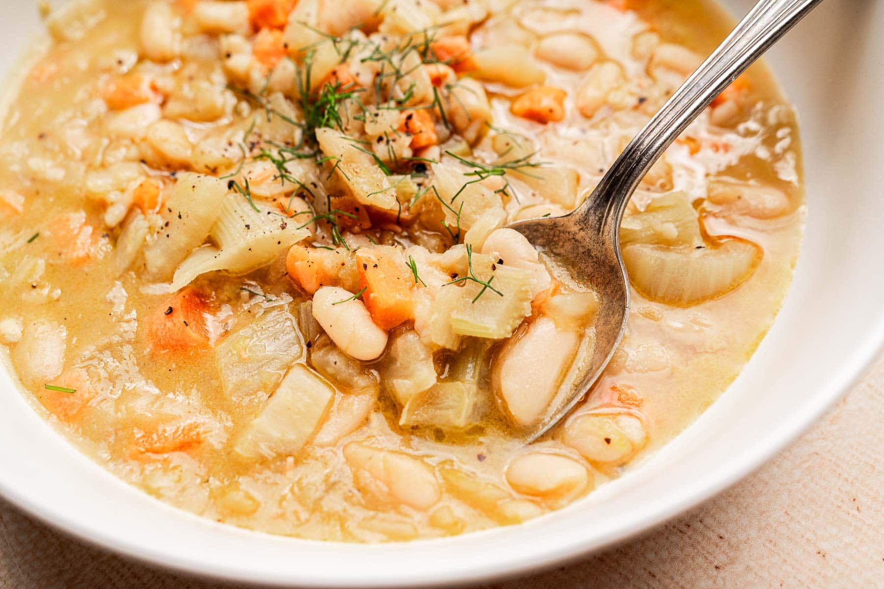 A bowl of Tuscan white bean soup with diced carrots, celery, and onions, garnished with herbs, with a spoon resting in the bowl.