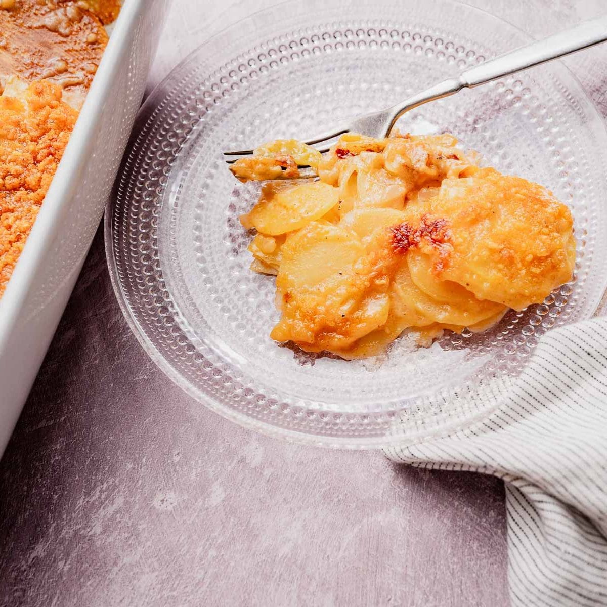A serving of cheesy potato casserole on a clear glass plate with a fork, next to a baking dish and a striped cloth.