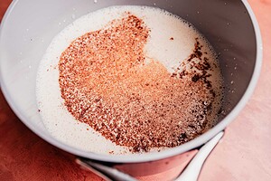 A saucepan containing milk with ground cinnamon and other spices sprinkled on top sits on a pink surface, ready to be transformed into Mexican hot chocolate.