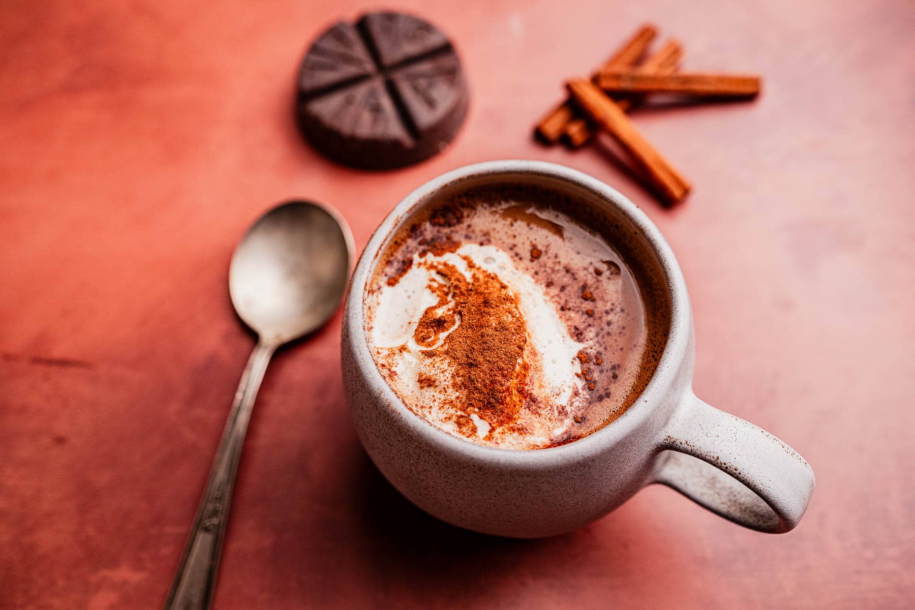 A mug of Mexican hot chocolate topped with cream and cinnamon sits on a reddish surface next to a spoon, chocolate disk, and cinnamon sticks.