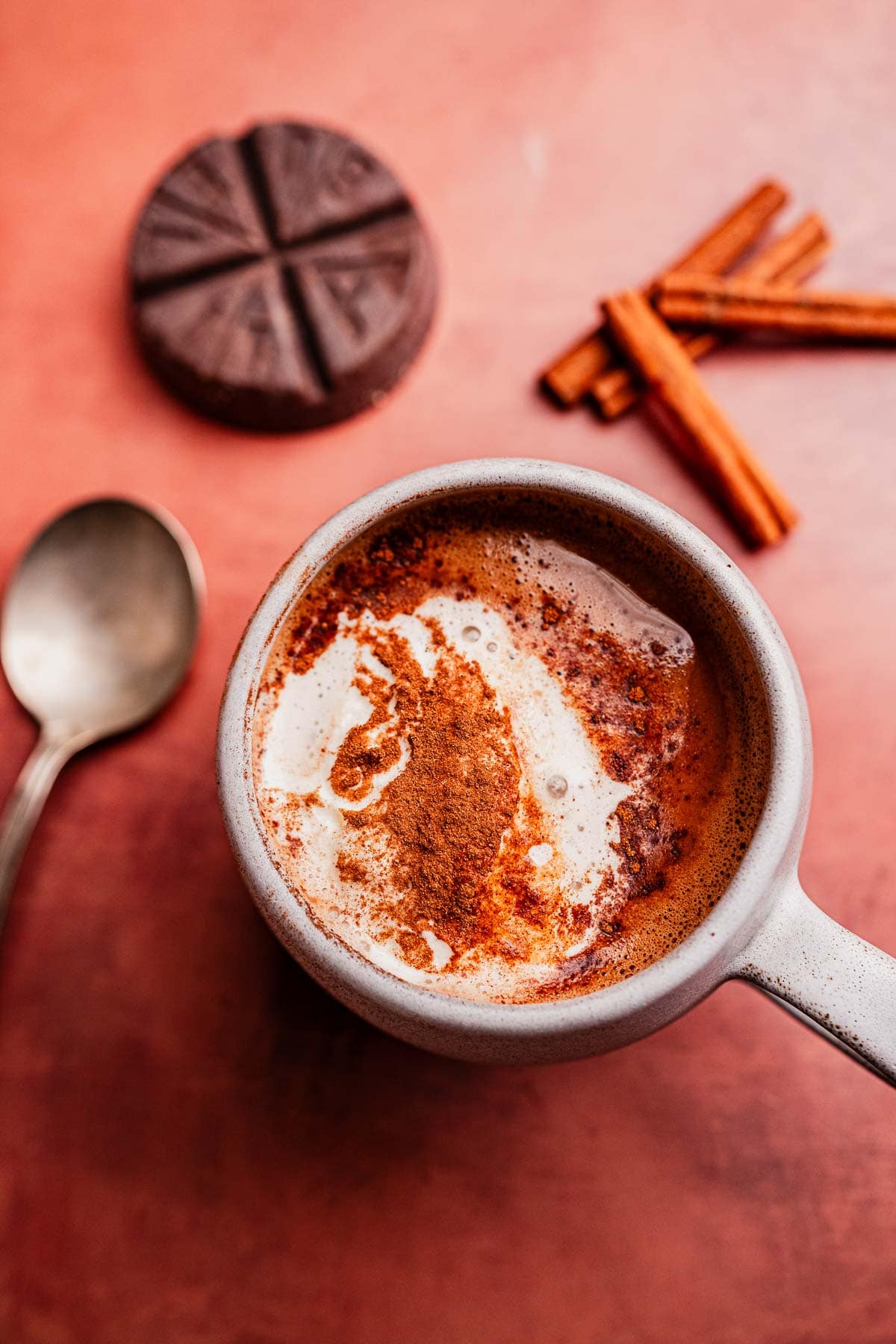 A mug of Mexican hot chocolate topped with cream and cinnamon sits on a pink surface next to a spoon, chocolate disc, and cinnamon sticks.