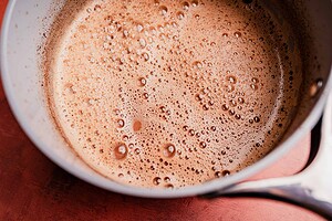 A close-up view of a saucepan containing frothy, bubbling Mexican hot chocolate mixture on a reddish-brown surface.