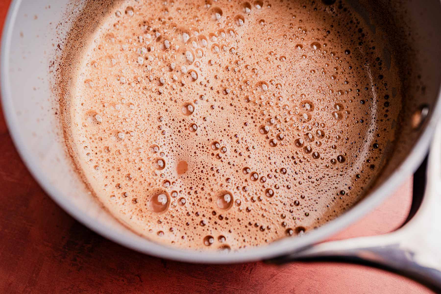 A close-up view of a saucepan containing frothy, bubbling Mexican hot chocolate mixture on a reddish-brown surface.