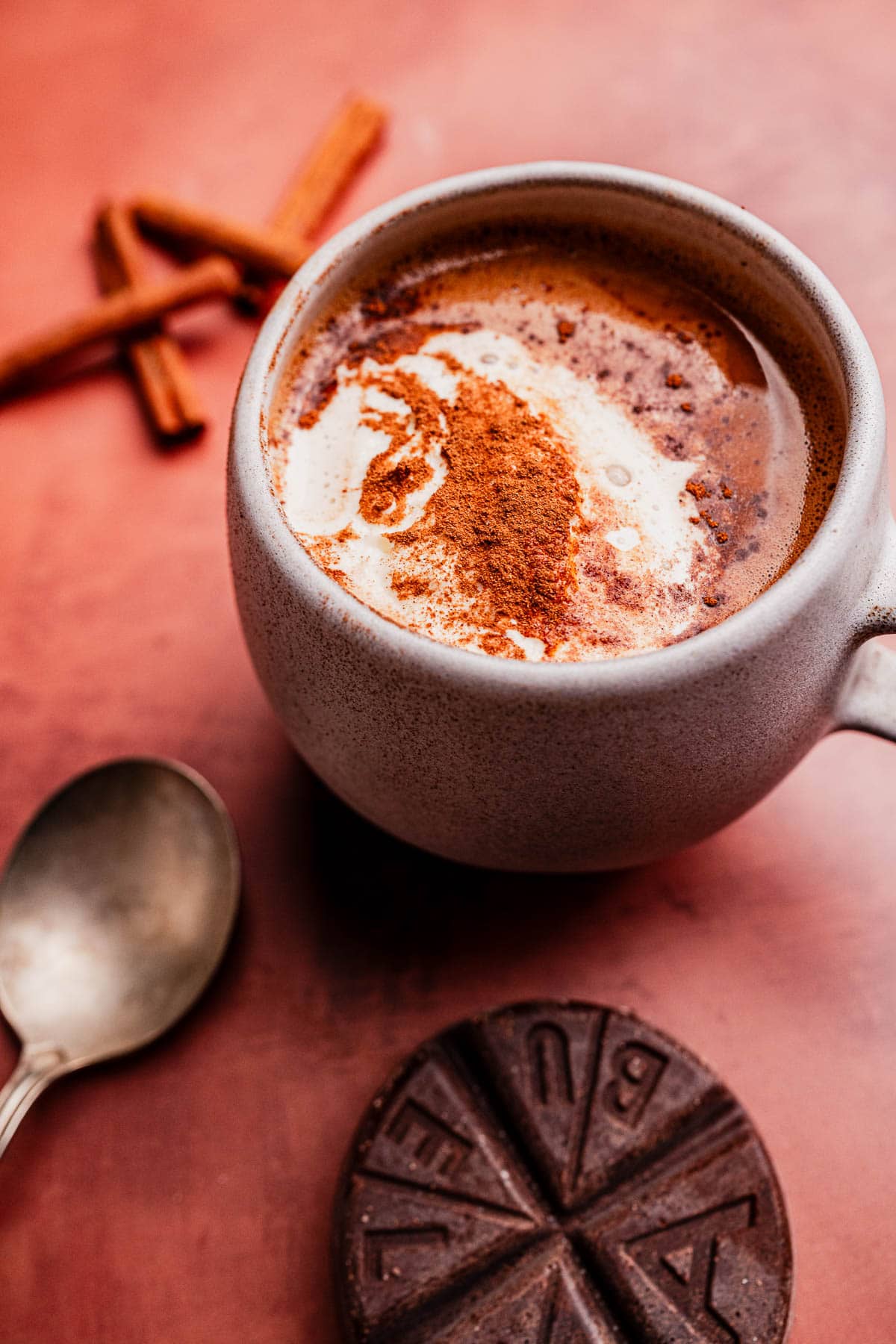 A mug of rich Mexican hot chocolate topped with cinnamon and cream, served with a spoon, cinnamon sticks, and a disk of chocolate on a pinkish surface.