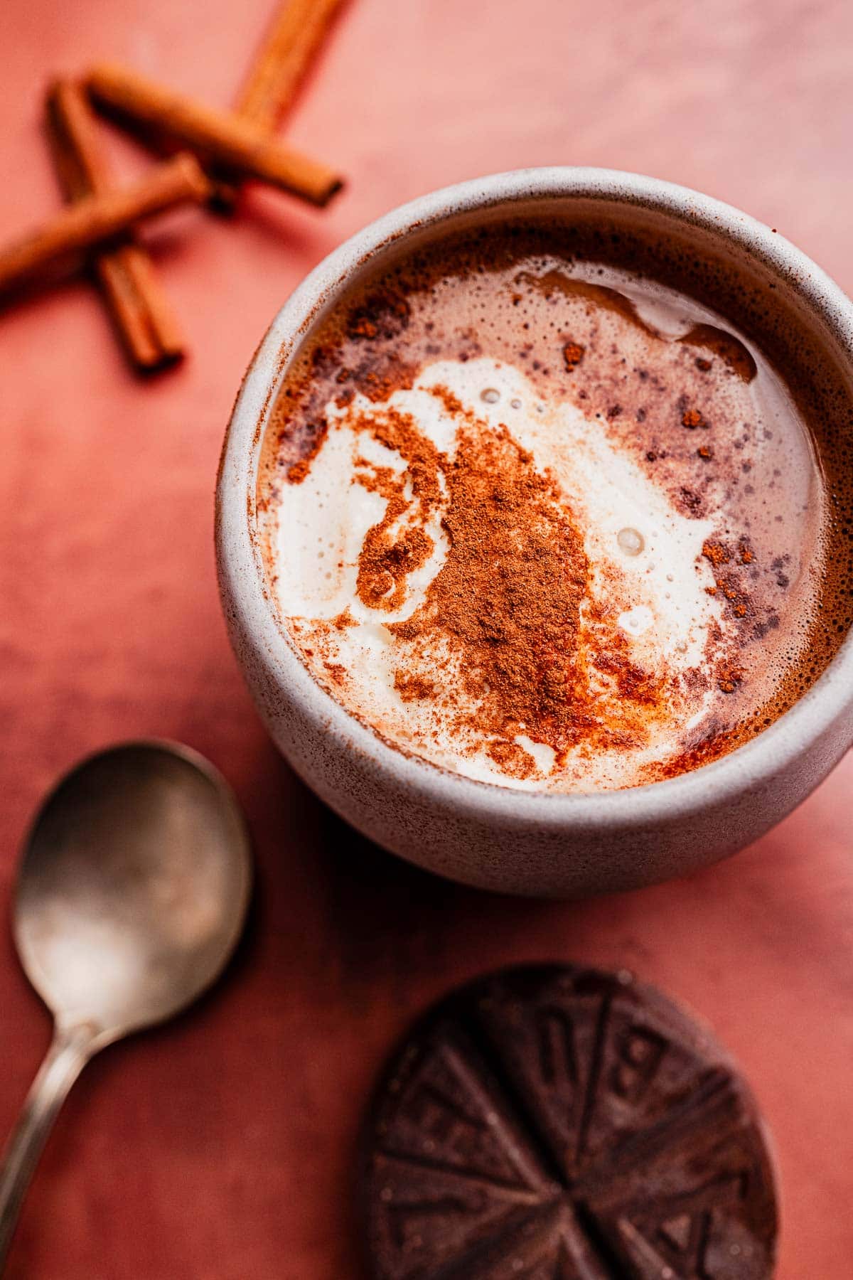 A ceramic mug of Mexican hot chocolate topped with cream and cinnamon sits on a pink surface beside a spoon, cinnamon sticks, and a round piece of chocolate.