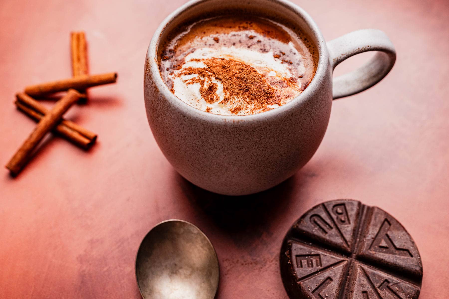 A mug of Mexican hot chocolate topped with cinnamon sits on a table with a spoon, cinnamon sticks, and a round tablet of Mexican chocolate nearby.