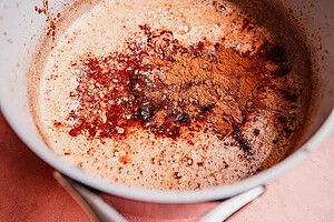 A saucepan containing a frothy liquid of Mexican hot chocolate, with cocoa powder and spices sprinkled on top.