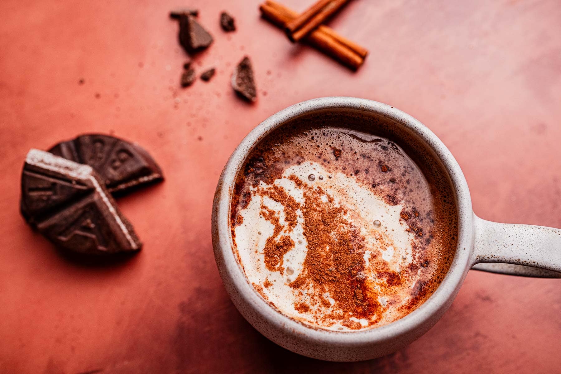 A mug of Mexican hot chocolate with cream and cinnamon, next to a round chocolate disk, broken pieces, and two cinnamon sticks on a reddish surface.
