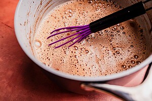 A saucepan filled with frothy Mexican hot chocolate mixture and a purple whisk resting inside, placed on a reddish-brown surface.