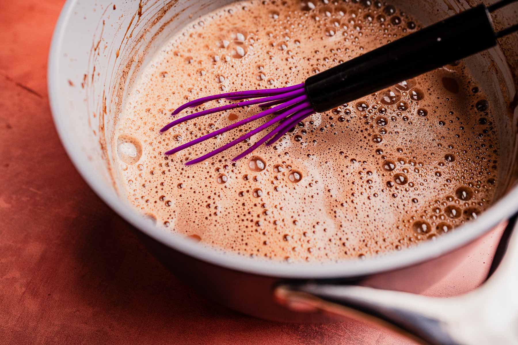 A saucepan filled with frothy Mexican hot chocolate mixture and a purple whisk resting inside, placed on a reddish-brown surface.