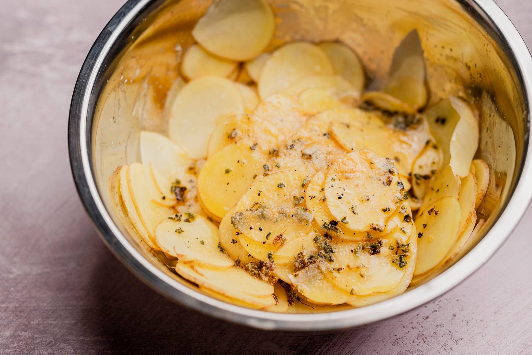 Thinly sliced potatoes in a metal bowl, seasoned with herbs, black pepper, and oil—perfect for making muffin tin potatoes, ready to be cooked.