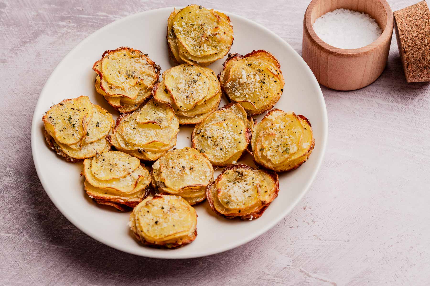 A white plate with stacks of muffin tin potatoes—baked potato slices sprinkled with herbs and seasoning—sits next to a small wooden bowl of coarse salt.